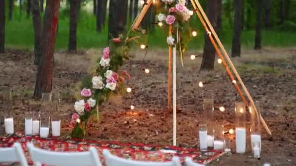 Bohemian tipi wooden arch decorated with pink roses, candles on carpet, pampass grass, wrapped in fairy lights on outdoor wedding ceremony venue in pine forest at night. Floristic compositions. — Stock Video