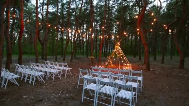 Ünnepi húros fények világítás boho tipi boltív dekoráció szabadtéri esküvői helyszín fenyves erdőben éjszaka. Vintage húr lámpák izzók koszorúk ragyog a székek felett a nyári vidéki esküvő. — Stock videók