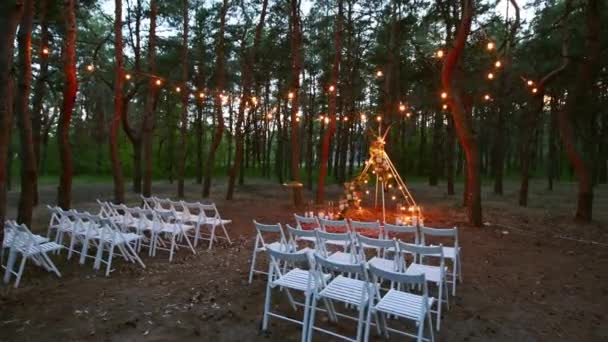 Çam ormanlarındaki açık hava düğün mekanında boho tipi kemer dekoru aydınlatması. Yazın kırsal düğünlerde sandalyelerin üzerinde parlayan eski sicim lambaları, çelenkler.. — Stok video