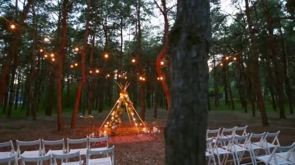 Festive lumières à cordes illumination sur boho tipi arche décor sur lieu de cérémonie de mariage en plein air dans la forêt de pins la nuit. Guirlandes d'ampoules à cordes vintage brillant au-dessus des chaises au mariage rural d'été. — Video