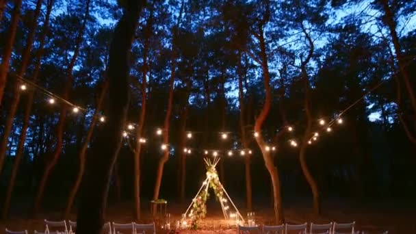 夜には松林の屋外結婚式会場にboho tipiアーチ装飾上の祭りの文字列照明。夏の田舎の結婚式で椅子の上に輝くヴィンテージストリングライト電球ガーランド. — ストック動画
