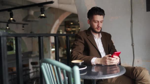Kafede oturup akıllı telefon kullanan şık, mutlu bir adam işaret veriyor ve gülümsüyor. Tabuttaki sosyal ağ uygulamasına bakan bir adam. Ofis çalışanı molada bir fincan kahveyle dinleniyor. İç mekan. — Stok video