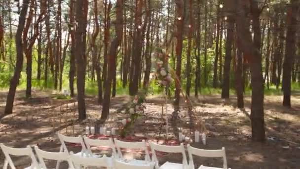Belle décoration bohème en arc de tipi sur le lieu de cérémonie de mariage en plein air dans la forêt de pins avec des cônes. Chaises, compositions floristiques de fleurs de roses, tapis, lumières de fées à cordes. Mariage rural d'été. — Video
