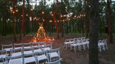 Çam ormanlarındaki açık hava düğün mekanında boho tipi kemer dekoru aydınlatması. Yazın kırsal düğünlerde sandalyelerin üzerinde parlayan eski sicim lambaları, çelenkler..