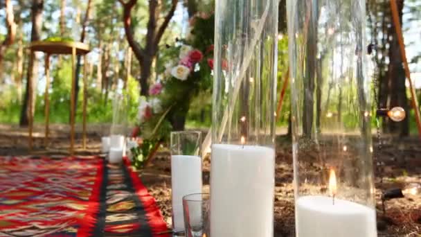 Velas queimando em vasos de vidro, frascos no tapete perto do arco de Tipi boêmio decorado com rosas e flores envoltas em luzes de fadas no local da cerimônia de casamento ao ar livre na floresta de pinheiros. Decoração rústica Boho. — Vídeo de Stock