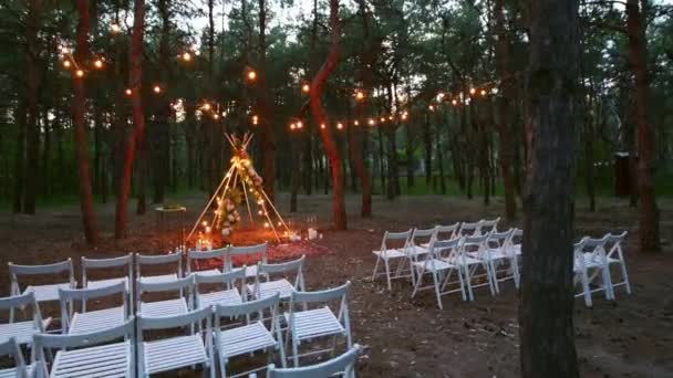 Çam ormanlarındaki açık hava düğün mekanında boho tipi kemer dekoru aydınlatması. Yazın kırsal düğünlerde sandalyelerin üzerinde parlayan eski sicim lambaları, çelenkler.. — Stok video