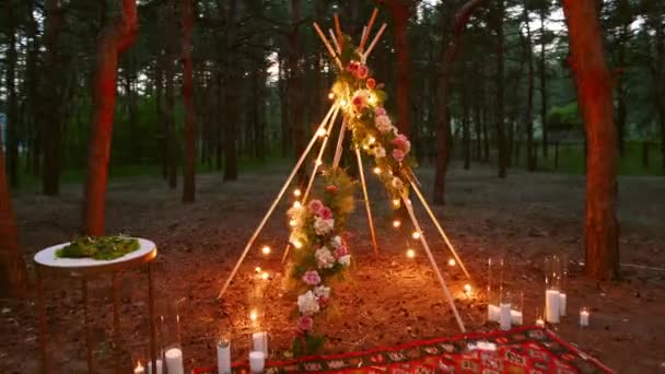 Boheemse tipi houten boog versierd met brandende kaarsen, rozen en pampass gras, verpakt in sprookjes verlichting op outdoor bruiloft locatie in dennenbos 's nachts. Bollen bloemenslinger schijnt. — Stockvideo