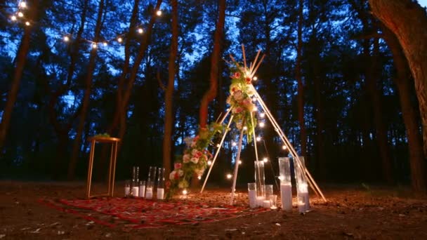 Luces festivas de cuerda iluminadas en la decoración del arco boho tipi en el lugar de la ceremonia de boda al aire libre en el bosque de pinos por la noche. Vintage luces de cadena bombilla guirnaldas que brillan por encima de las sillas en verano boda rural. — Vídeo de stock