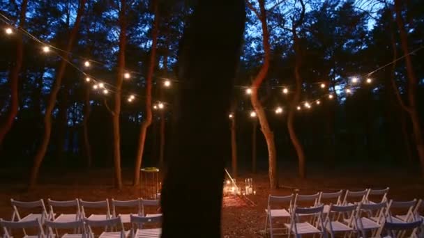 Festlig sträng ljus belysning på boho tipi båge inredning på utomhus bröllop plats i tallskog på natten. Vintage sträng lampor glödlampa girlanger lyser ovan stolar på sommaren lantligt bröllop. — Stockvideo