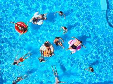 Havuzda, şişme flamingo, kuğu ve şiltelerle parti veren arkadaşlar. Mutlu gençler güneşli bir günde lüks bir tatil köyünde dinleniyorlar. Yukarıdan bak. Bikinili kızlar güneşte güneşleniyor..