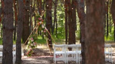 Çam ormanındaki açık hava düğün mekanında konili güzel bir bohem çadır kemeri süslemesi. Sandalyeler, çiçek kompozisyonları güller, halı, ip perisi ışıkları. Yaz kırsal düğünü.