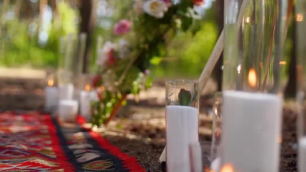 Kaarsen branden in glazen vazen, flessen op tapijt in de buurt van Boheemse tipi boog versierd met rozen en bloemen gewikkeld in feeënlichtjes op outdoor bruiloft locatie in dennenbos. Boho rustieke inrichting. — Stockvideo