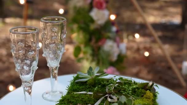 Copas de vino, anillos de boda ropa de cama de hierba y musgo en mesa blanca junto al arco de tipi bohemio decorado en estilo boho con flores y velas y sillas blancas para invitados. — Vídeos de Stock