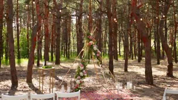 Prachtige Boheemse tipi boog decoratie op outdoor bruiloft locatie in dennenbos met kegels. Stoelen, bloemige bloemcomposities van rozen, tapijt, string fee lichten. Zomer landelijke bruiloft. — Stockvideo
