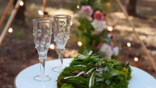 Copas de vino, anillos de boda ropa de cama de hierba y musgo en mesa blanca junto al arco de tipi bohemio decorado en estilo boho con flores y velas y sillas blancas para invitados. — Vídeos de Stock