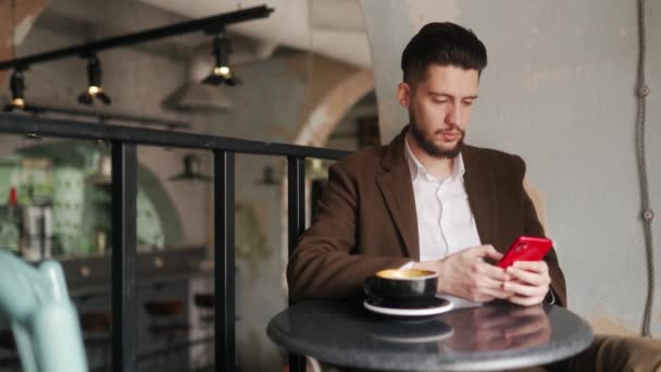 Joven con clase usando teléfono inteligente sentado en la cafetería. Chico navegando aplicación de red social en coffeshop. Trabajador de oficina descansando en el descanso con una taza de café. Escena interior. — Vídeos de Stock