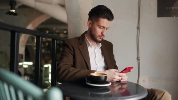 カフェに座っているスマートフォンを使用してクラスの若い男。コーヒーショップでソーシャルネットワークアプリを閲覧する男。オフィスワーカーはコーヒーを飲みながら休憩している。屋内シーン. — ストック動画