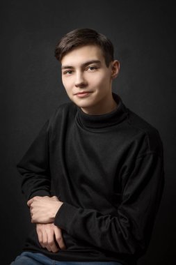 Siyah arka planda siyah elbiseli, kahverengi gözlü, koyu saçlı, gülümseyen yakışıklı genç bir adamın portresi; bir sandalyede oturuyor; doğrudan kamera lensine bakın;