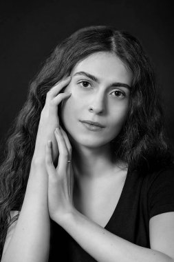 Siyah arka planda siyah elbiseli, kahverengi saçlı ve kahverengi gözlü güzel bir kadının siyah beyaz portresi. Yüzüne dokunan ellerle doğrudan kameranın lensine bakın.