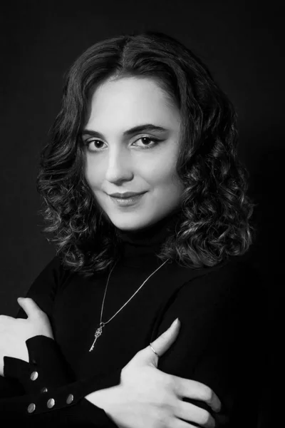 black and white portrait of a smiling beautiful young woman with curly hair and black eyes  with hands, in black clothes on a black background; look directly into the camera lens; vertical