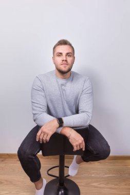Gri kazaklı ve koyu kotlu genç sakallı bir adam beyaz bir duvara karşı çapraz kollarıyla bir bar taburesinde oturuyor. Düşünceli bir ifadeyle kameraya bakıyor..