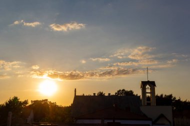 Güneş, çan kulesi olan bir köy kilisesinin arkasında batıyor ve manzaraya sıcak bir ışık tutuyor. Gün sona ererken sakin bir atmosfer ortamı kaplıyor..