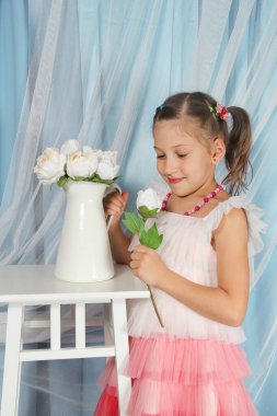 Beyaz bir elbise ve pembe etek giyen genç bir kız beyaz güllerle süslenmiş bir masanın yanında duruyor. Yumuşak bir gülümsemeyle çiçeklere bakar, mavi dokulu bir perdenin arka planına bakar..