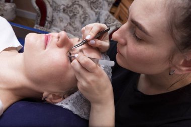 Genç bir kadın, güzellik salonunda profesyonel bir güzellik uzmanı tarafından kirpiklerini büyütüyor. Güzellik uzmanı cımbızla kadınların doğal kirpiklerini dikkatlice uzatıyor..