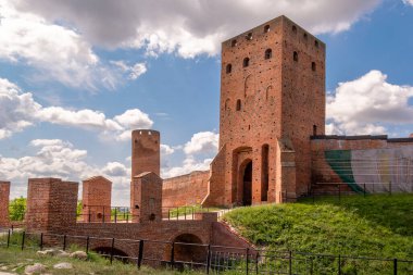 Yeşil bir alanda uzun bir tuğla kule ve eski duvarlar görülür. Gökyüzünde birçok bulut var. Ziyaretçiler kuleye yakın yürüyorlar. Bu sitenin tarihi bir önemi var ve bölge manzarası sunuyor..