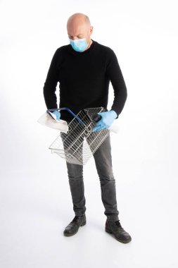 Ağız korumalı ve eldivenli bir adam beyaz arka planda izole edilmiş bir alışveriş sepetini temizliyor.