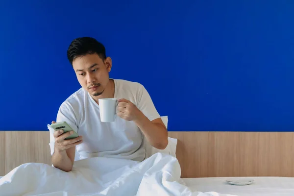 Pria dengan smartphone dan kopi di tempat tidur dengan latar belakang ruang fotokopi biru. — Stok Foto