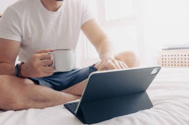 Adam yatak odasında tablet ve kahveyle çalışıyor. Evden çalışma kavramı.
