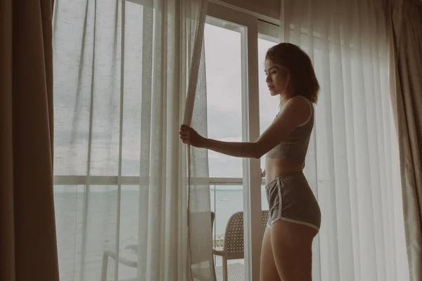 Atractiva mujer mira la vista detrás de la cortina en el hotel. —  Fotos de Stock