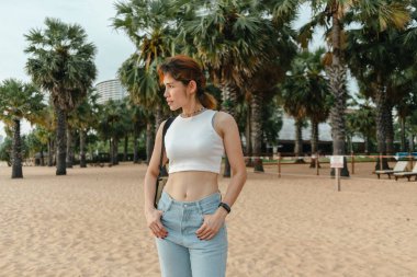 Arka planda palmiye ağaçları olan bir kadın turist sahilde yürüyor..