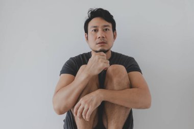 Siyah elbiseli Taylandlı erkek, beyaz bir stüdyonun zemininde çıplak ayakla oturuyor. Dizleri çekilmiş, öne doğru eğik, etkileyici ve hassas bir duruş sergiliyor. Sakin, minimalist bir iç mekanda içsel duygular olduğunu gösteriyor..