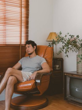 Rahat Taylandlı erkek rahat bir evde, pencerenin yanındaki klasik deri koltukta, huzurlu bir şekilde sakin bir anın tadını çıkarıyor.