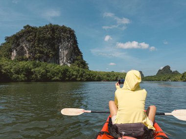 Sarı kapüşonlu biri kireçtaşı adasına doğru kayak yapıyor, açık yeşil su, dramatik uçurumlar, parlak gökyüzü, gevşek keşif