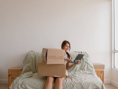 Taylandlı bir kadın, yatak odasında, Asya kökenli bir tabletle yatakta otururken odak ile teslimat kutusunu inceliyor.