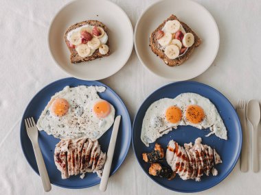 İki mavi tabak kızarmış yumurta ve pilav, iki kase muzlu tost ve yabanmersini ızgara tavuk dilimleri, çatal bıçak takımı ve keten masa üstü, sıcak doğal ışık, samimi bir çift.