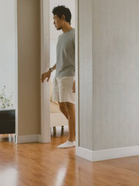 Tarafsız ifadeli genç Taylandlı adam, güneşli, ahşap bir dairede, yumuşak bej bir duvarın yanından geçiyor, sakin bir yaşam tarzı ve huzurlu bir ortam sunuyor..