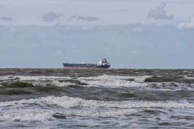 Yazın güneşli bir günde Cadzand yakınlarındaki Northsea sahilinin köpük başlarıyla dalgakıranlarda bir kargo gemisi.