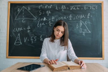 Genç bir öğrenci elinde bir kitapla masada oturuyor, arka planda geometri yazıtları olan bir karatahta. Bilgileri tablet ve kitapla kontrol ederiz..