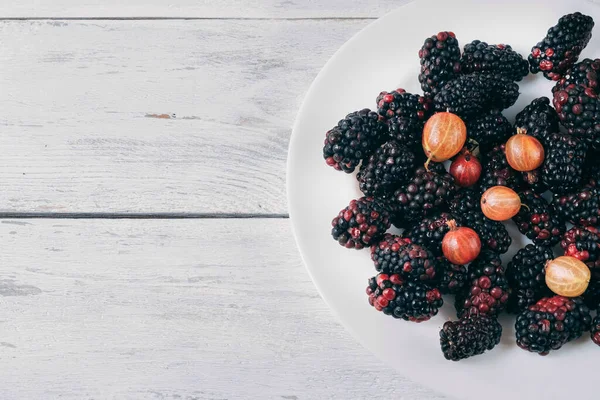 Berry mix of gooseberries and blackberries agrus on a white plate on a wooden background. Plate with berries half frame on the right side. View from above. Copy space