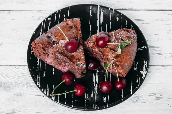 Vişneli siyah bir tabakta iki parça browni. Böğürtlenlerle süslenmiş beyaz ahşap bir masanın üzerinde pasta tabağı..