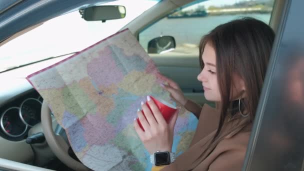 Une fille est assise dans le siège du conducteur d'une voiture avec une carte papier et boit du café — Video