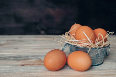 Süslü, paslanmaz çelik bir leğende, ahşap bir masada dekoratif saman üzerine tavuk yumurtası. Bir kasede üç yumurta, masada iki tane daha. Uzayı kopyala Metnin altına yerleştir.