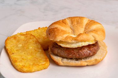   fried egg  with sausage patty  on a croissant served with hash browns