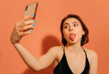 Genç bir kadın selfie çekiyor ve turuncu arka planda dil çıkararak yüzünü buruşturuyor.