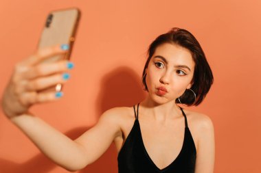 Genç bir kadın selfie çekiyor ve turuncu arka planda dudaklarını buruşturuyor.