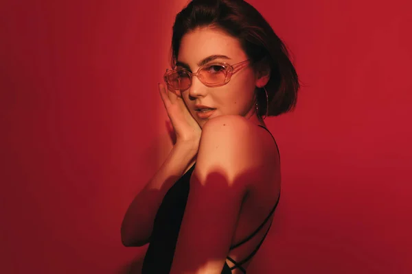 Siyah kolsuz tişörtlü ve güneş gözlüklü, kırmızı arka planda kameraya bakan utangaç genç bir kadın.